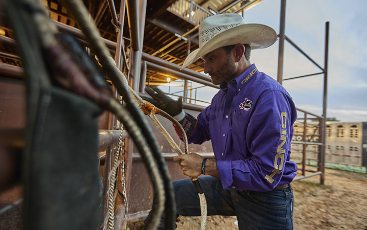 Man wearing a purple shirt, cowboy hat, and a glove getting ready to ride a bull by warming up the Rossin on his bull rope.